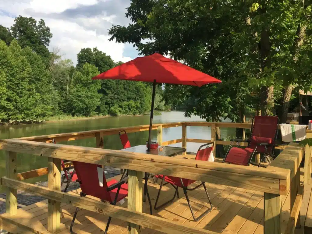 Wooden deck overlooking a calm river with red patio chairs, glass table, and a large red umbrella surrounded by trees.